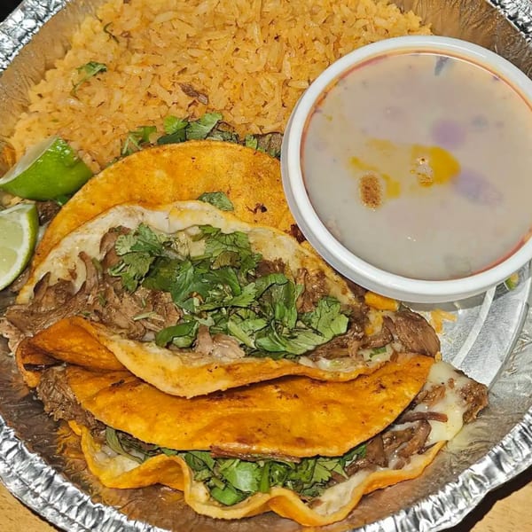 Three birria tacos with cilantro, rice, lime, and a side of consommé in a foil container.