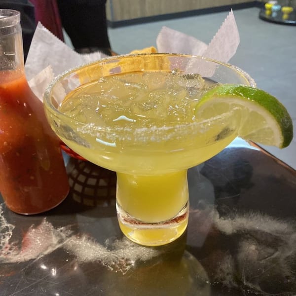 A vibrant margarita with a salted rim and lime wedge, next to a bottle of red hot sauce.
