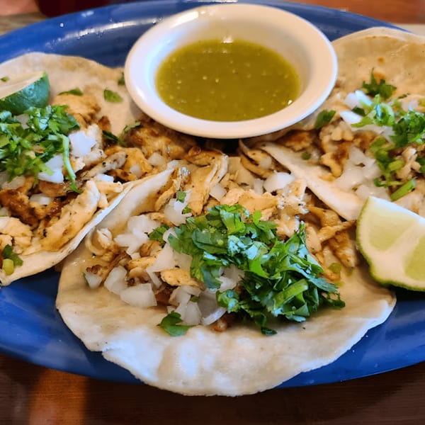 Three chicken tacos with cilantro, onions, and lime wedges, served with green salsa on a blue plate.