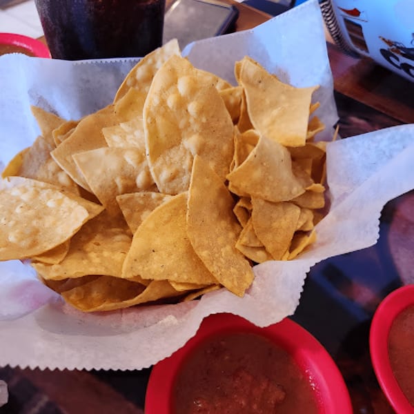 A basket of Los Compas' tortilla chips with two red bowls of salsa and a drink.