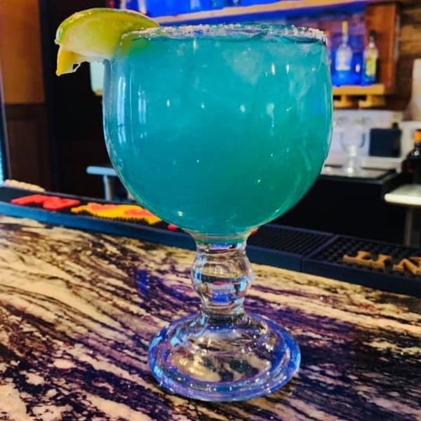 A large blue margarita with a lime wedge and salted rim sits on a marble bar.