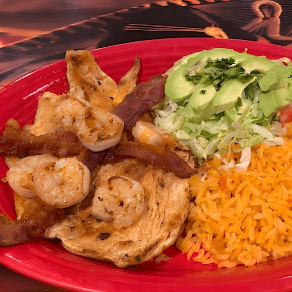 A vibrant red plate holds a Mexican dish with grilled chicken, shrimp, bacon, rice, and avocado.