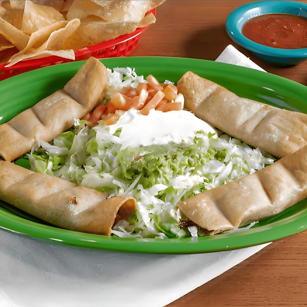 Four taquitos arranged around a bed of shredded lettuce, sour cream, and diced tomatoes on a green plate.